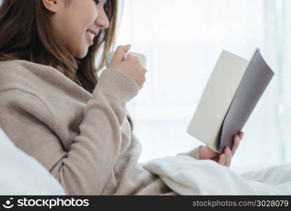 Beautiful asian woman enjoying warm coffee and reading book on b. Beautiful asian woman enjoying warm coffee and reading book on bed in her bedroom.Asia female wearing comfortable sweater holding a book and cup of coffee.lifestyle asia woman at home concept.