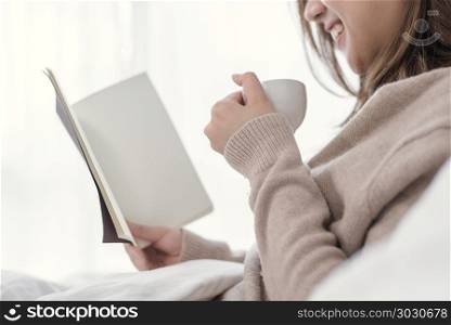Beautiful asian woman enjoying warm coffee and reading book on b. Beautiful asian woman enjoying warm coffee and reading book on bed in her bedroom.Asia female wearing comfortable sweater holding a book and cup of coffee.lifestyle asia woman at home concept.
