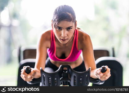 Beautiful Asian woman exercising on stationary cycling machine in indoor fitness gym, determination face. Sport recreational activity, people workout, or healthy lifestyle concept