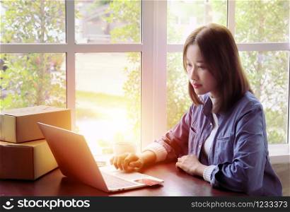 Beautiful Asian woman in casual using laptop technology for running small business start up marketing with cardboard box logistic delivery. young entrepreneur using credit card shopping payment,