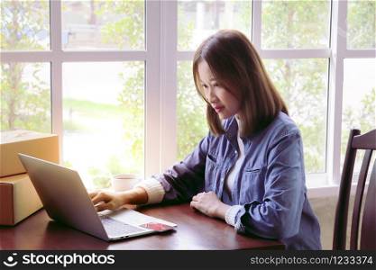 Beautiful Asian woman in casual using laptop technology for running small business start up marketing with cardboard box logistic delivery. young entrepreneur using credit card shopping payment,