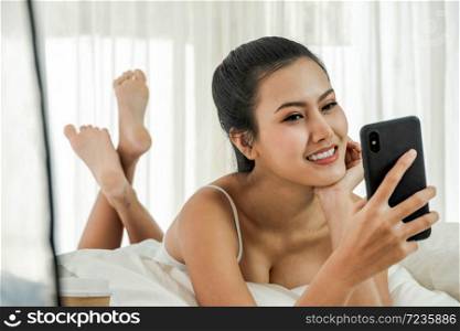 Beautiful asian woman just woke up and laying on the bed using mobile phone and texing or video call in the morning light. Cell Phone, Gadget, Technology concept. Copy Space.