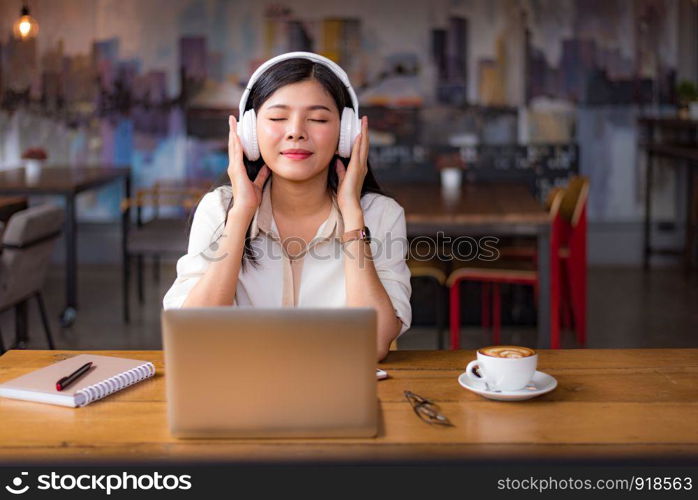 Beautiful Asian woman relaxing and listening to music in cafe with laptop computer and coffee cup. People and lifestyles concept. Freelance happy workplace theme. University and college theme.