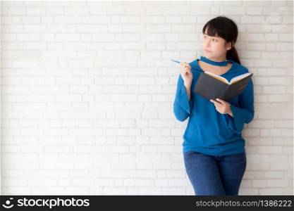 Beautiful asian woman smiling standing thinking and writing notebook on concrete cement white background at home, girl homework on book, education and lifestyle concept.
