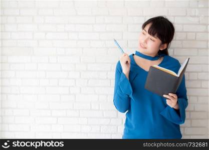Beautiful asian woman smiling standing thinking and writing notebook on concrete cement white background at home, girl homework on book, education and lifestyle concept.