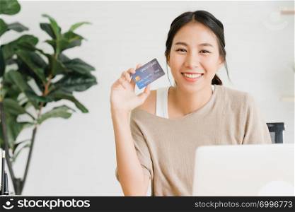Beautiful Asian woman using computer or laptop buying online shopping by credit card while wear sweater sitting on desk in living room at home. Lifestyle woman at home concept.