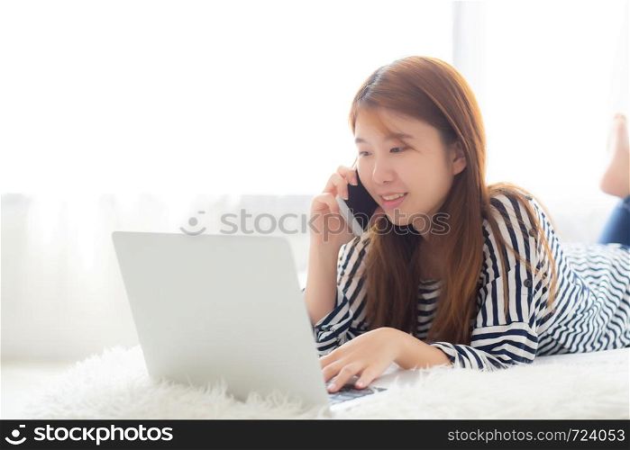 Beautiful asian young woman lying on bed using laptop and talking mobile smart phone at bedroom for leisure and relax, freelance with girl working notebook, communication concept.