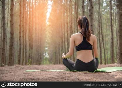 Beautiful Asian young woman lying on green mat and doing yoga in forest. Exercise and meditation concept. Peaceful and countryside concept. Pine wood in summer theme. Back view
