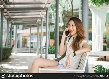Beautiful Asian young woman using smartphone for talking, reading and texting sitting at bench in urban city in summer and sunny day. Lifestyle women travel and relax in the city concept.