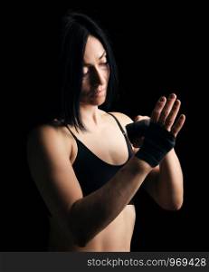 beautiful athletic girl with black hair rewinds her hand with a black elastic bandage before training, dark background