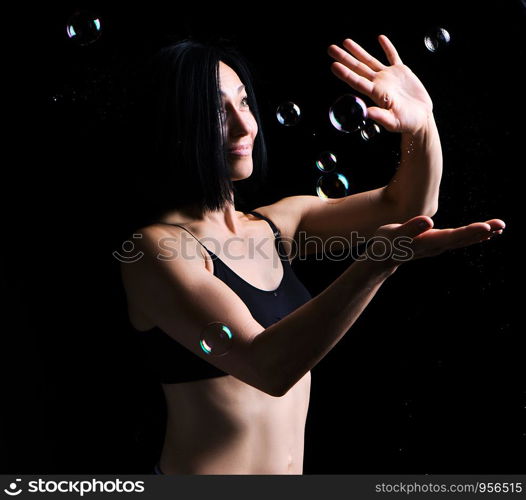 beautiful athletic woman with muscular body dressed with a black topic catches soap bubbles on a black background, studio portraits in a dark key