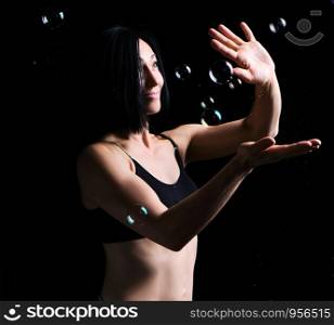 beautiful athletic woman with muscular body dressed with a black topic catches soap bubbles on a black background, studio portraits in a dark key