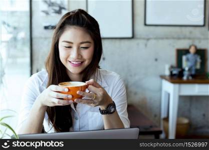 Beautiful Attractive Asian Business woman wearing white shirt smile and holding coffee in Coffee shop cafe with computer laptop on the table feeling so happiness and comfortable