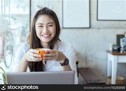 Beautiful Attractive Asian Business woman wearing white shirt smile and holding coffee in Coffee shop cafe with computer laptop on the table feeling so happiness and comfortable