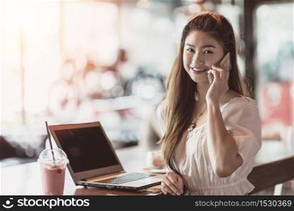 Beautiful Attractive Asian woman working with laptop smile and talking with customer in smartphone to get idea and requirement for success job outside office in coffee shop,Business Concept,Warm Tone