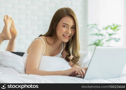 Beautiful Attractive Asian young woman lying on bed and using computer laptop for using social media and shopping online relax in cozy bedroom in holiday,Lifestyle in home Concept 