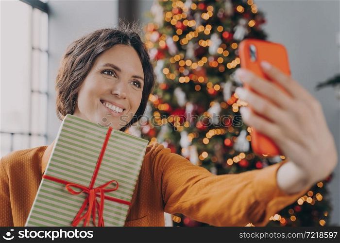 Beautiful attractive brunette female taking selfie with Christmas gift box near decorated xmas tree at home, cheerful european woman holding New Year present and making self photo on smartphone. Beautiful brunette female taking selfie with Christmas gift box near decorated xmas tree at home