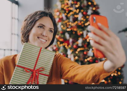 Beautiful attractive brunette female taking selfie with Christmas gift box near decorated xmas tree at home, cheerful european woman holding New Year present and making self photo on smartphone. Beautiful brunette female taking selfie with Christmas gift box near decorated xmas tree at home