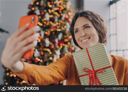 Beautiful attractive brunette female taking selfie with Christmas gift box near decorated xmas tree at home, cheerful european woman holding New Year present and making self photo on smartphone. Beautiful brunette female taking selfie with Christmas gift box near decorated xmas tree at home