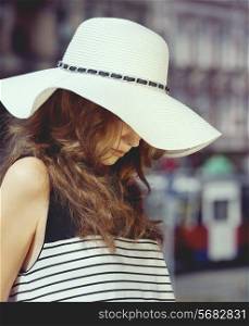 Beautiful, attractive woman in summer hat on the street in summer day. She has got brown, curly, long hair and she is wearing stripped cloth.