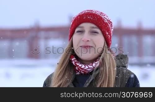 Beautiful attractive woman with deep blue eyes standing outdoors in the snowfall and looking at camera. Smiling young woman enjoying snow shower in wintertime.