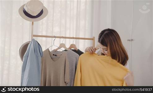 Beautiful attractive young Asian woman choosing her fashion outfit clothes in closet at home or store. Girl think what to wear sweater. Home wardrobe or clothing shop changing room.