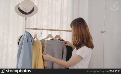 Beautiful attractive young Asian woman choosing her fashion outfit clothes in closet at home or store. Girl think what to wear sweater. Home wardrobe or clothing shop changing room.