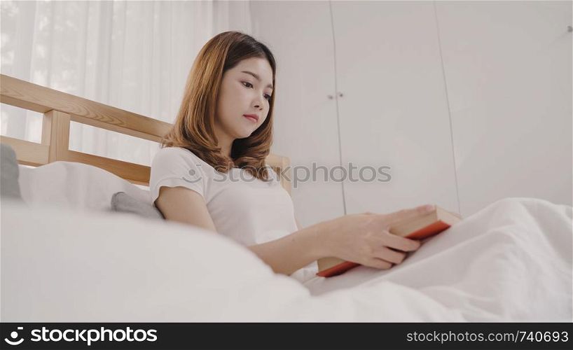 Beautiful attractive young Asian woman reading a book while lying on the bed when relax in her bedroom at home in the morning. Lifestyle women using relax time at home concept.