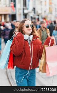 Beautiful attractive young girl in jumper and jeans with sunglasses and bags walks at a crowded street after shopping
