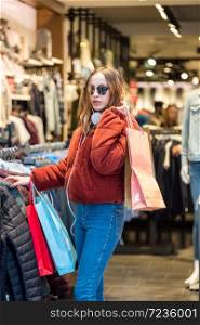 Beautiful attractive young girl in jumper with headphone and sunglasses chooses clothes thinking, looking at clothes while shopping in store