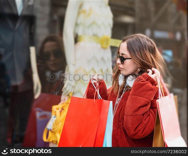 Beautiful attractive young girl in jumper with headphone and sunglasses looks at clothes display windows at shopping store
