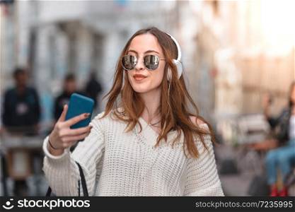 Beautiful attractive young trendy girl in jumper with headphones smartphone and sunglasses listens music while walking in crowd