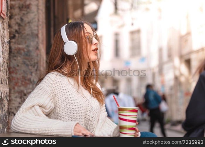 Beautiful attractive young trendy girl with headphones and sunglasses sits and listens music at a street cafe while drinking fruit juice