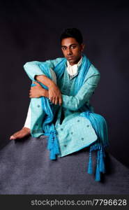 Beautiful authentic Indian hindu man in typical ethnic groom attire sitting relaxed on top of rock leaning on knee. Bangali male wearing a light blue agua decorated Dhoti with shawl.