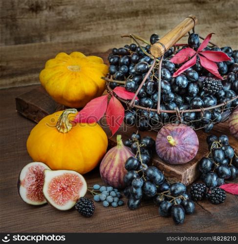 Beautiful autumn still life: black grapes in a basket on the old cutting board as well as ripe figs, red raspberries, orange pumpkin and blackberries with autumn leaves on the wooden background