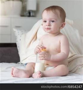 Beautiful baby boy in diapers sitting on bed and holding his bottle