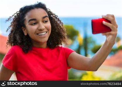 Beautiful biracial mixed race African American teenager teen girl young woman smiling with perfect teeth taking selfie on cell phone outside by the sea in summer