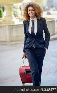 Beautiful black woman smiling and carrying a rolling suitcase in urban background Businesswoman wearing suit with trousers and tie, afro hairstyle.