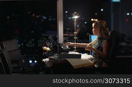 Beautiful blond business woman working overtime at night in executive office, answering phone call and talking on wired telephone. City lights are visible in background from a large window