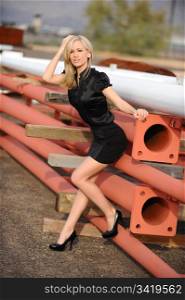 Beautiful blond fashion model, wearing a short black skirt, black shirt and back stilettos leaning against materials at a construction site outdoors.