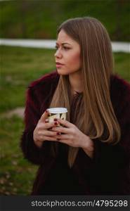 Beautiful blond girl with fur coat drinking a cofee in the park