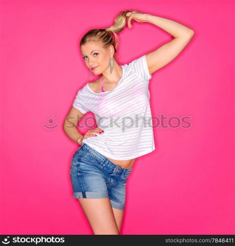 Beautiful blond woman wearing a white shirt and a denim shorts and posing against a pink studio background