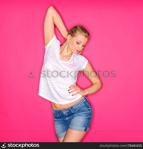 Beautiful blond woman wearing a white shirt and a denim shorts and posing against a pink studio background