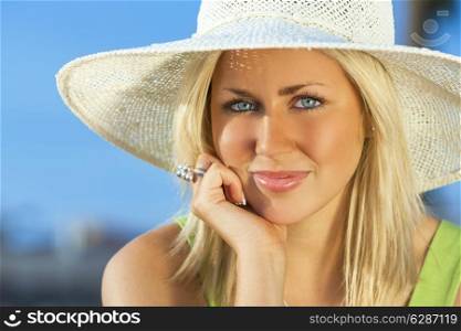 Beautiful blond young woman or girl in her twenties happy smiling wearing sun hat resting on her hand in sunshine on summer day with blue sky