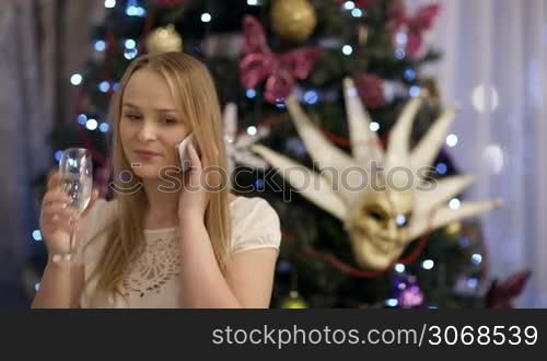 Beautiful blond young woman smiling and drinking champagne while talking on mobile phone, with a Christmas tree decorated with lights, baubles, bows and a Venetian mask in the background