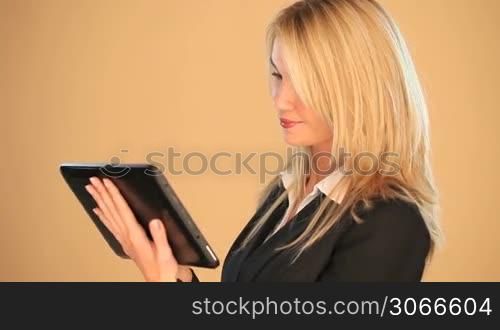 Beautiful blonde businesswoman working on a tablet looking up with a friendly smile against a brown background