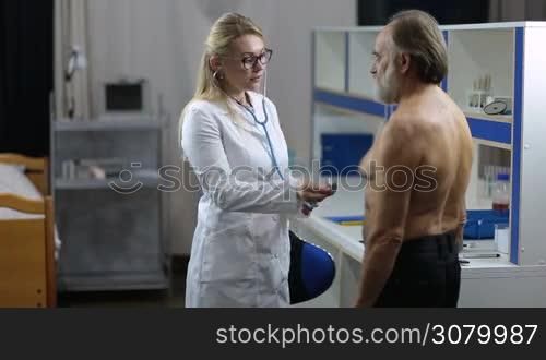 Beautiful blonde female physician in eyeglasses and lab coat using stethoscope to exam the heart of an ill senior male patient in medical office. Female doctor with stethoscope auscultating heart rate of elderly man during medical checkup.