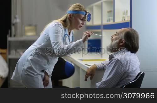 Beautiful blonde female physician in lab coat examining senior man&acute;s throat with tongue depressor at clinic. Young female ENT specialist checking elderly male patient&acute;s throat using a tongue depressor and head mirror to look inside mouth and tonsils.