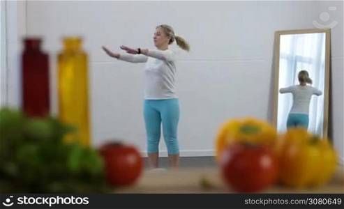 Beautiful blonde female stretching her arms and waming up before exercising at home with blurred fresh vegetables on foreground. Healthy young woman doing fitness exercises in domestic room.