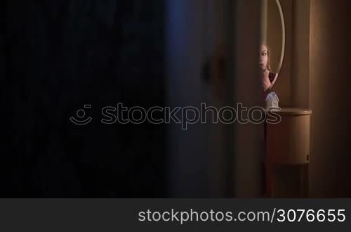 Beautiful blonde girl applying lipstick with a brush in front of the mirror. Reflection of young pretty lady in the mirror using red lipstick. Woman sitting on table with Christmas decorations and doing makeup preparing to celebrate Christmas.
